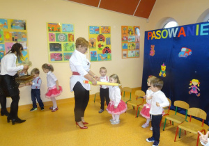 Pani dyrektor Maria Królikowska trzyma w ręku dużą żółtą kredkę, którą przykłada dziewczynce do lewego ramienia.